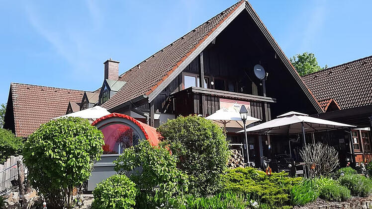 Gem&uuml;tliches Gasthaus mit Holzdach, Sonnenschirmen und &uuml;ppiger Bepflanzung. Idyllische Atmosph&auml;re, ideal f&uuml;r eine Pause auf einem fr&auml;nkischen Campingplatz.