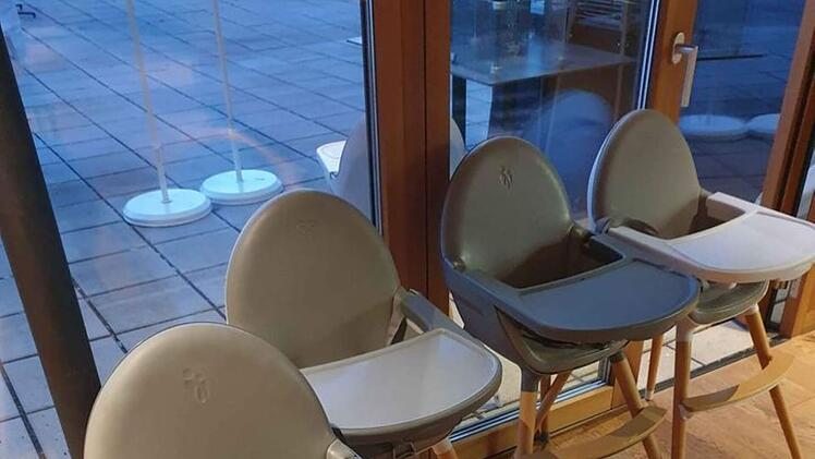 Das Bild zeigt vier graue Kinderstühle mit Tabletts in einem Raum mit Holzfußboden und großen Fenstern. Links stehen dekorative Gläser.
