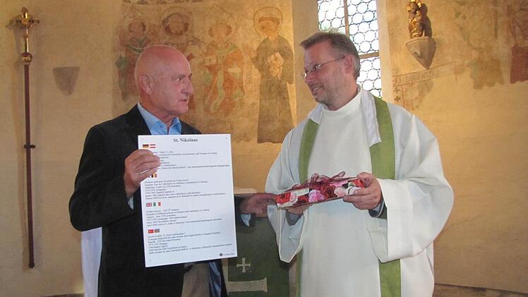 Das Gemeindeglied Jürgen Wérobel La Rochelle hatte als Begrüßungsgeschenk für Pfarrer Hans-Jürgen Pöschl eine neue Wandtafel mit den historischen Daten der Nikolauskapelle dabei. Foto: Martin Koch