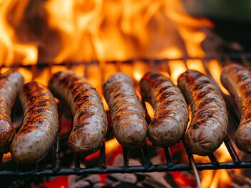 Bratwürste liegen auf einem Grillrost. Dahinter flammt Feuer auf. Im Artikel geht es um einen großen Bratwurst- beziehungsweise Grillwurst-Test.