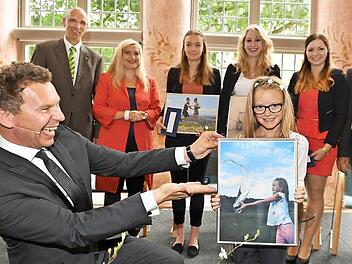 Moderator Roman Roell präsentiert das Siegerbild mit dessen Motiv. Hinten rahmen MGO-Geschäftsführer Walter Schweinsberg (l.) mit Gesundheitsministerin Melanie Huml und Rainer Drewello (Pfleger) die Preisträgerinnen Pauline Greb, Celine Eckert und Lea Örtelt (v.r.) ein.
