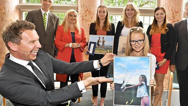 Moderator Roman Roell präsentiert das Siegerbild mit dessen Motiv. Hinten rahmen MGO-Geschäftsführer Walter Schweinsberg (l.) mit Gesundheitsministerin Melanie Huml und Rainer Drewello (Pfleger) die Preisträgerinnen Pauline Greb, Celine Eckert und Lea Örtelt (v.r.) ein.