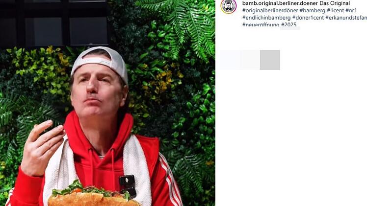 Ein Mann in rotem Hoodie und weißer Kappe isst genussvoll ein großes Sandwich. Hintergrund mit grünen Pflanzen. Überraschungsbesuch eines Kult-Comedians zur Eröffnung eines Döner-Ladens.