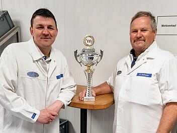 Zwei M&auml;nner in wei&szlig;en Kitteln pr&auml;sentieren einen gro&szlig;en Pokal. Sie wirken stolz, m&ouml;glicherweise auf ihre Leistung beim Metzgercup 2026, wo fr&auml;nkische Metzgereien ausgezeichnet wurden.