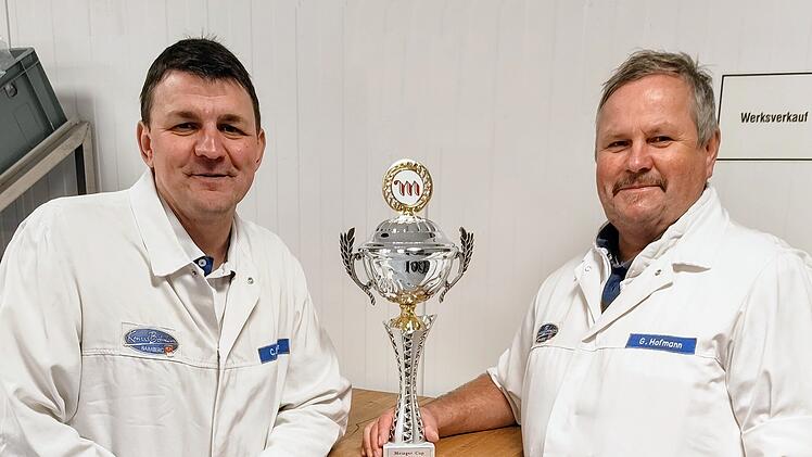 Zwei M&auml;nner in wei&szlig;en Kitteln pr&auml;sentieren einen gro&szlig;en Pokal. Sie wirken stolz, m&ouml;glicherweise auf ihre Leistung beim Metzgercup 2026, wo fr&auml;nkische Metzgereien ausgezeichnet wurden.