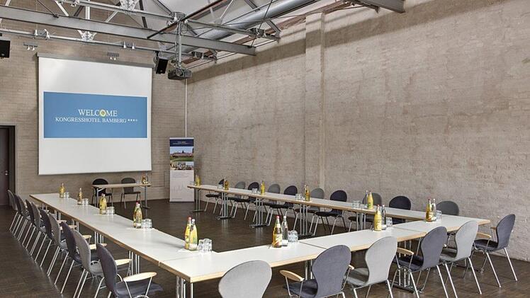 Moderner Konferenzraum in Bamberg, U-förmige Tischanordnung, komfortable graue Stühle, Wasserflaschen und Gläser bereit, großer Bildschirm mit Begrüßung für Kongresshotel Bamberg angezeigt.