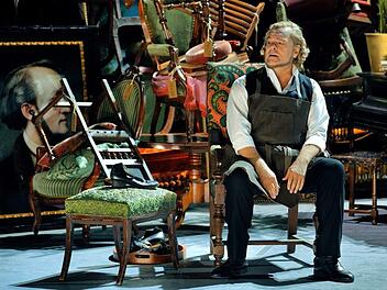 Der Bariton-Sänger Michael Volle als Hans Sachs in der Schusterstube des 2. Akts, hinter der sich das Wahnfried-Mobiliar des 1. Akts als Konkursmasse auftürmt.  Bayreuther Festspiele/Enrico Nawrath