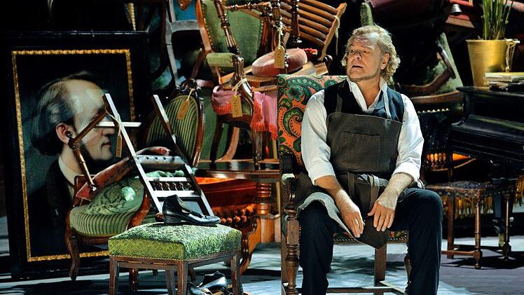 Der Bariton-Sänger Michael Volle als Hans Sachs in der Schusterstube des 2. Akts, hinter der sich das Wahnfried-Mobiliar des 1. Akts als Konkursmasse auftürmt.  Bayreuther Festspiele/Enrico Nawrath