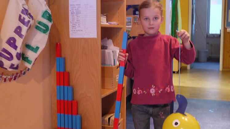 Cosma zeigt die blauroten Holzstäbe zum Einprägen von Längen. Handfeger und Schaufel daneben gehören auch zu Montessoris Material.