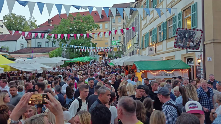 Das Bild zeigt ein gut besuchtes Volksfest in einer Altstadt mit bunten Wimpeln. St&auml;nde und Menschen f&uuml;llen die Stra&szlig;e, die Stimmung ist lebhaft und fr&ouml;hlich.