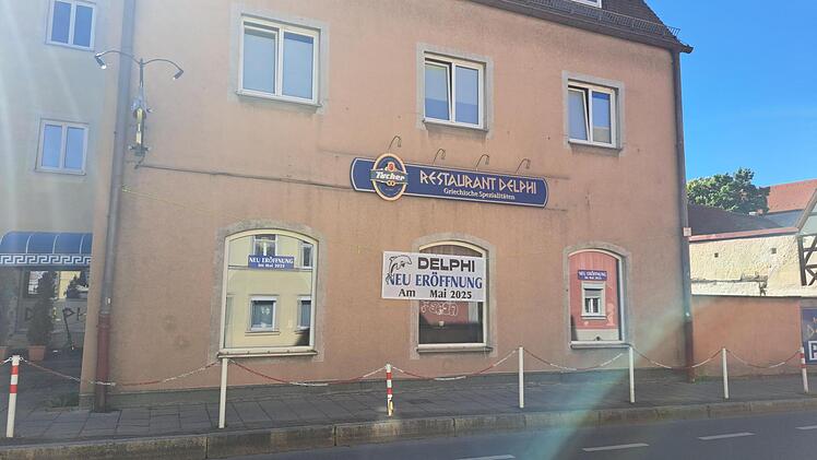 Das Foto zeigt das griechische Restaurant "Delphi" mit Ankündigungen zur Neueröffnung im Mai 2025. Die Fassade ist beige, das Logo blau, und Schilder informieren über die Rückkehr.