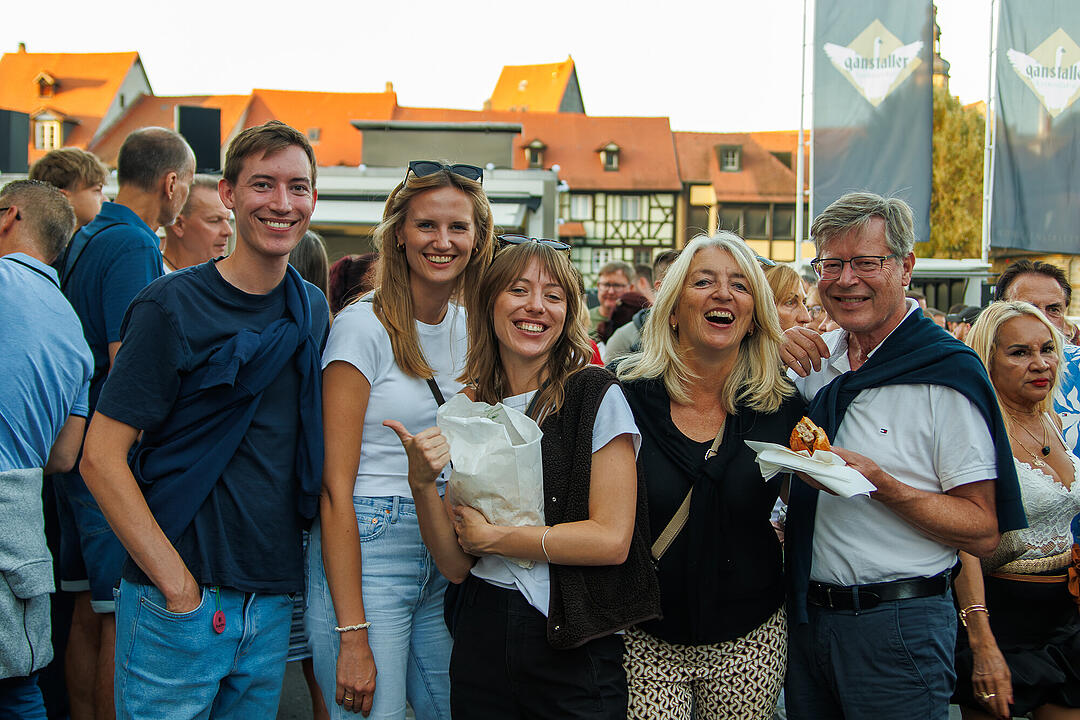 Eine fr&ouml;hliche Gruppe von Menschen l&auml;chelt bei der Bamberger Sandkerwa 2026. Sie genie&szlig;en das Fest bei gutem Wetter und entspannter Stimmung vor traditionellen Fachwerkh&auml;usern.