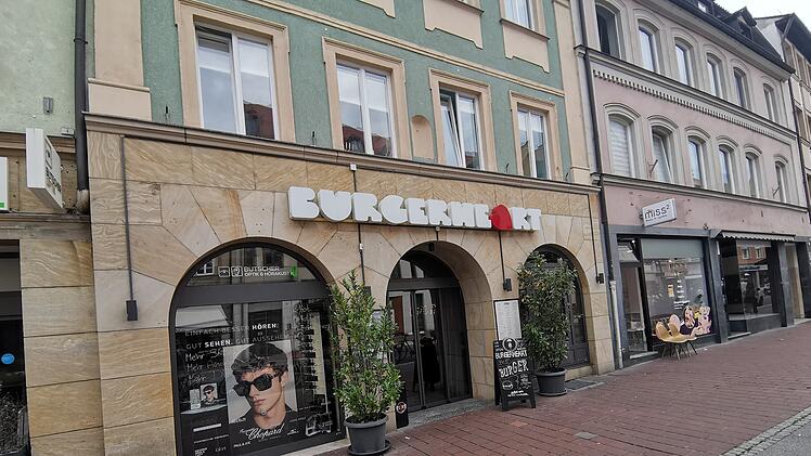Ein Restaurant der fränkischen Burger-Kette ist abgebildet. Das Logo zeigt teilweise verdeckte Buchstaben, die Fassade ist beige und grün, daneben sind Geschäfte zu sehen.