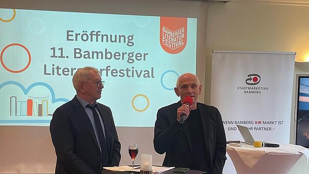 Zwei M&auml;nner stehen auf einer B&uuml;hne bei der Er&ouml;ffnung des 11. Bamberger Literaturfestivals. Ein Mann spricht in ein Mikrofon. Im Hintergrund ist eine Festivalpr&auml;sentation zu sehen.