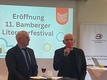 Zwei M&auml;nner stehen auf einer B&uuml;hne bei der Er&ouml;ffnung des 11. Bamberger Literaturfestivals. Ein Mann spricht in ein Mikrofon. Im Hintergrund ist eine Festivalpr&auml;sentation zu sehen.