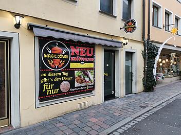 Das Bild zeigt die Fassade des neuen "Magic Döner" in Bamberg. Ein großes Schild kündigt die Neueröffnung mit einer 1-Cent-Aktion an. Die Straße ist gepflastert, mit Läden daneben.