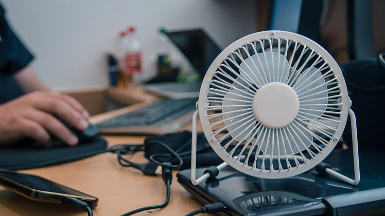 A small desktop USB powered fan or ventilator to cool the earea on a work desk with desktop computer. Hands of a person seen working in the background.