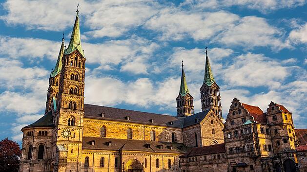 Der Bamberger Dom im Sonnenschein mit seinen markanten T&uuml;rmen vor einem teilweise bew&ouml;lkten Himmel. Historische Architektur im Vordergrund.