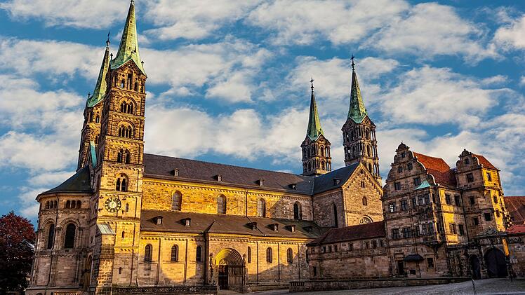 Der Bamberger Dom im Sonnenschein mit seinen markanten T&uuml;rmen vor einem teilweise bew&ouml;lkten Himmel. Historische Architektur im Vordergrund.