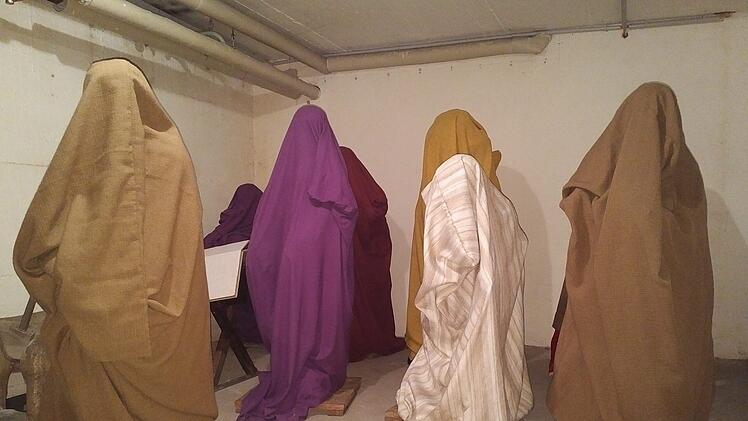 Verschiedene in Stoff gehüllte Figuren stehen in einem kahlen Raum. Sie sind in Braun, Violett und anderen Erdtönen gekleidet. Der Zweck ist unklar, es wirkt wie eine Installation.