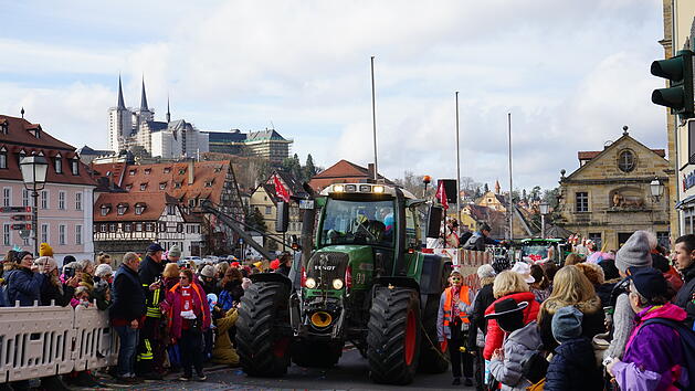 Ein Faschingsumzug mit geschm&uuml;ckten Traktoren zieht durch eine Stadt mit Fachwerkh&auml;usern. Viele Menschen in Kost&uuml;men s&auml;umen die Stra&szlig;en. Im Hintergrund sind Kircht&uuml;rme zu sehen.