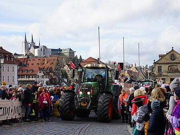 Ein Faschingsumzug mit geschmückten Traktoren zieht durch eine Stadt mit Fachwerkhäusern. Viele Menschen in Kostümen säumen die Straßen. Im Hintergrund sind Kirchtürme zu sehen.