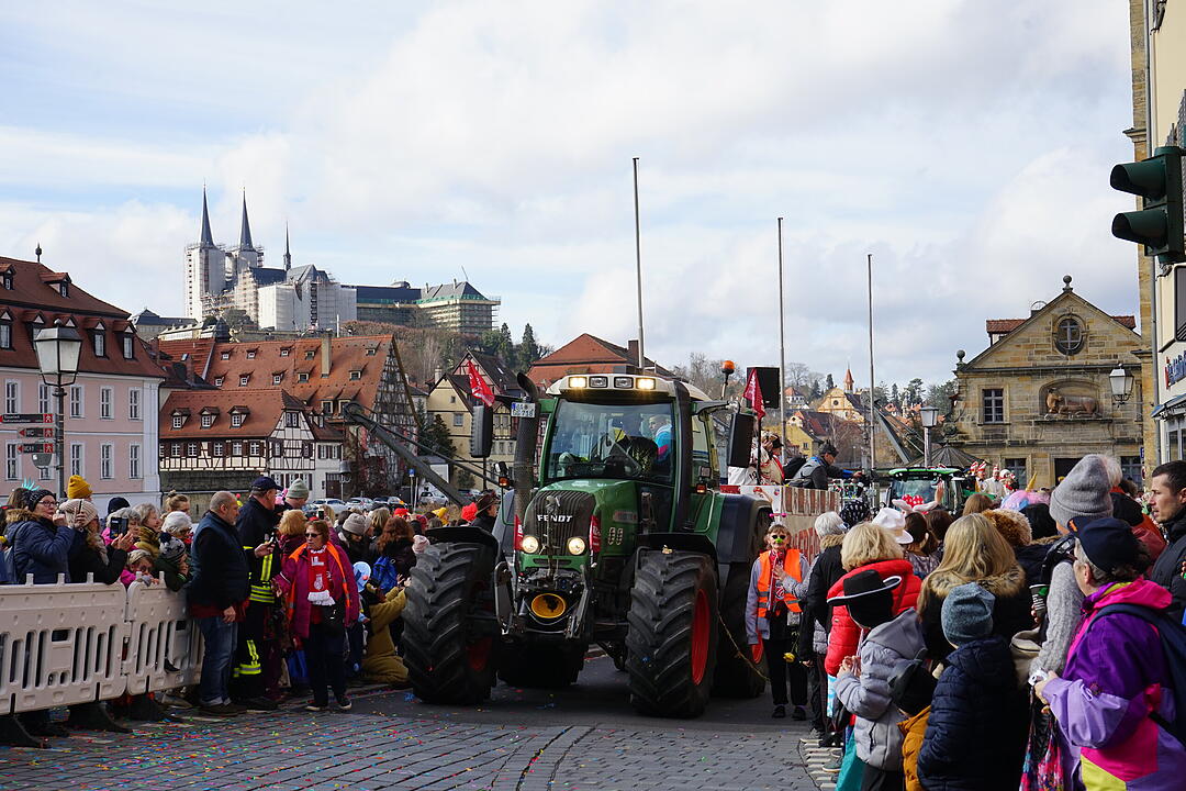 Ein Faschingsumzug mit geschmückten Traktoren zieht durch eine Stadt mit Fachwerkhäusern. Viele Menschen in Kostümen säumen die Straßen. Im Hintergrund sind Kirchtürme zu sehen.