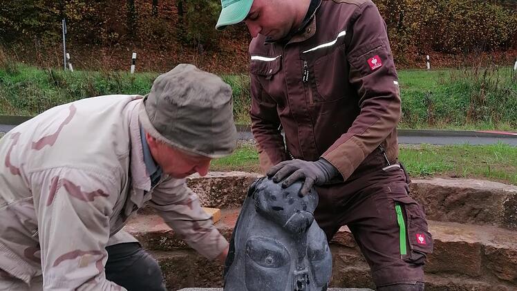 Die Gemeindearbeiter Berthold Schaab und Michael Detsch befestigen den von Gerwin Kellermann gemeißelten Rucksack aus Basaltstein.