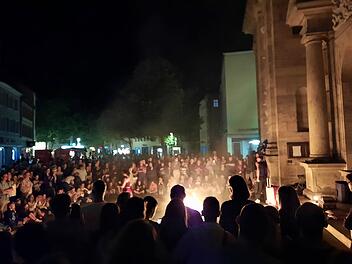 Eine gro&szlig;e Menschenmenge beobachtet eine n&auml;chtliche Feuershow vor einer historischen Geb&auml;udekulisse, die von warmem, dramatischem Licht beleuchtet wird.