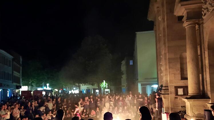 Eine große Menschenmenge beobachtet eine nächtliche Feuershow vor einer historischen Gebäudekulisse, die von warmem, dramatischem Licht beleuchtet wird.