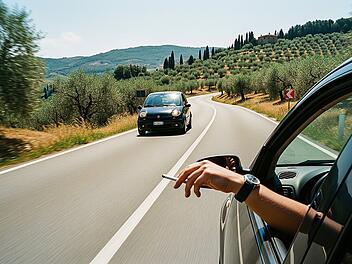 Eine italienische Landstraße, irgendwo zwischen malerischen Hügeln und Olivenhainen, aufgenommen in Italien im Sommer 2025. Im Vordergrund fährt ein dunkler Kleinwagen mit heruntergelassenem Fenster; eine linke Hand wirft gerade einen Zigarettenstummel aus dem Fenster.