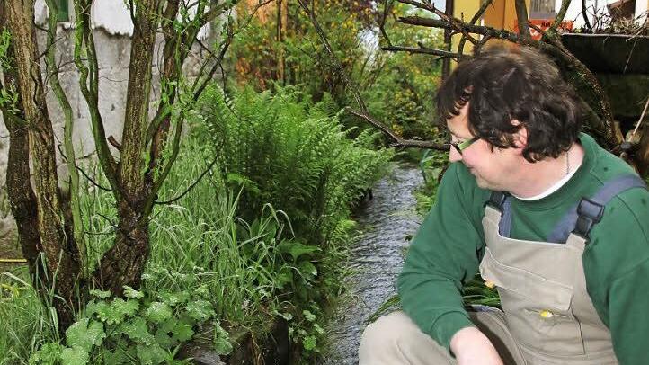 Michael Franz blickt auf den Bach, der sich durch seinen Garten schlängelt. 2002 trat dieser über die Ufer, das Wasser stand so hoch, wie der Putz an der Hauswand (links im Bild) abgetragen ist. Foto: Corinna Igler