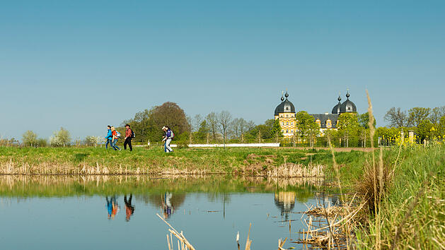 Wanderer genie&szlig;en den Fr&uuml;hling in der Fr&auml;nkischen Toskana. Im Hintergrund ist ein pr&auml;chtiges Geb&auml;ude, das sich im ruhigen Wasser eines Sees spiegelt. B&auml;ume und klarer Himmel umrahmen die Szene.