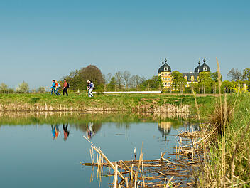 Wanderer genie&szlig;en den Fr&uuml;hling in der Fr&auml;nkischen Toskana. Im Hintergrund ist ein pr&auml;chtiges Geb&auml;ude, das sich im ruhigen Wasser eines Sees spiegelt. B&auml;ume und klarer Himmel umrahmen die Szene.