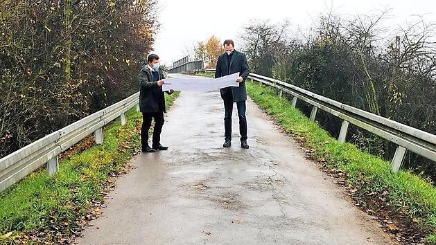 Die alte Br&uuml;cke &uuml;ber den Bahngleisen soll nach dem Willen der Bahn abgerissen und durch ein neues Br&uuml;ckenbauwerk ersetzt werden, um den alten Bahn&uuml;bergang "Weide" auflassen zu k&ouml;nnen. B&uuml;rgermeister Carsten Joneitis und Bauamtsleiter Markus Klarmann setzen sich vor Ort mit der Planung auseinander. Foto: Gemeinde