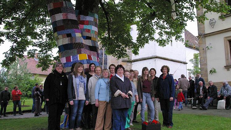 Rund um die Dorflinde präsentiert sich Gemünda im neuen Gewand. "Wir sind die Guerilla-Gruppe der Kirchengemeinde", scherzte Pfarrerin Kathrin Neeb (links) bei der Präsentation des bestrickten Dorfplatzes am Sonntag. Diese Art der Straßenkunst wird als Urban oder Guerilla Knitting bezeichnet. Rund 15 Frauen der Gemünner Lichtstube unter Leitung von Barbara Hämmelmann (rechts vorne) haben das Projekt auf die Beine gestellt. Foto: Bettina Knauth