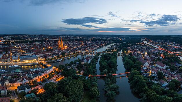 Panorama der Stadt Regensburg in Bayern mit dem Fluss Donau dem beleuchtetem Dom und der steinernen Brücke im Sommer zur blauen Stunde im Zwielicht, Deutschland