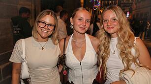 Drei junge Frauen l&auml;cheln in die Kamera bei der Bamberger Sandkerwa 2026. Die Stra&szlig;e ist belebt und festlich beleuchtet.