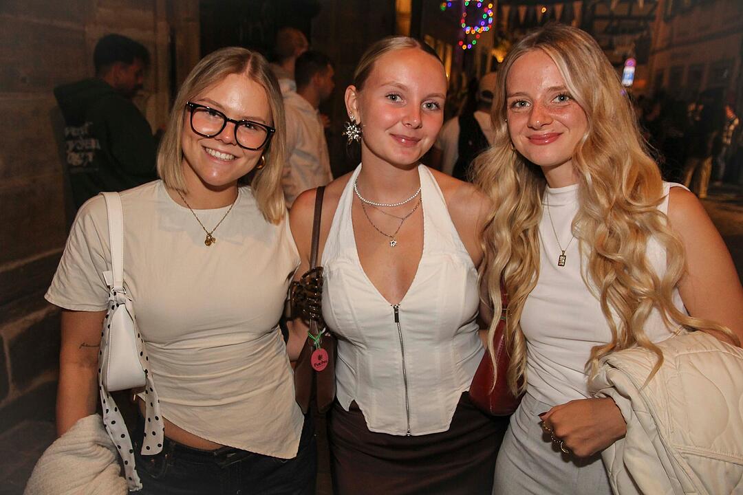 Drei junge Frauen l&auml;cheln in die Kamera bei der Bamberger Sandkerwa 2026. Die Stra&szlig;e ist belebt und festlich beleuchtet.