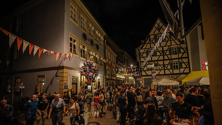 Menschenmenge feiert abends in einer geschm&uuml;ckten Altstadtstra&szlig;e w&auml;hrend eines Volksfests. Fahnen, Luftballons und Fachwerkh&auml;user schaffen eine festliche Atmosph&auml;re.