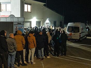 Menschen stehen nachts in einer langen Schlange vor einem beleuchteten Geb&auml;ude. Ein Kleintransporter parkt in der N&auml;he. Sie tragen warme Kleidung.
