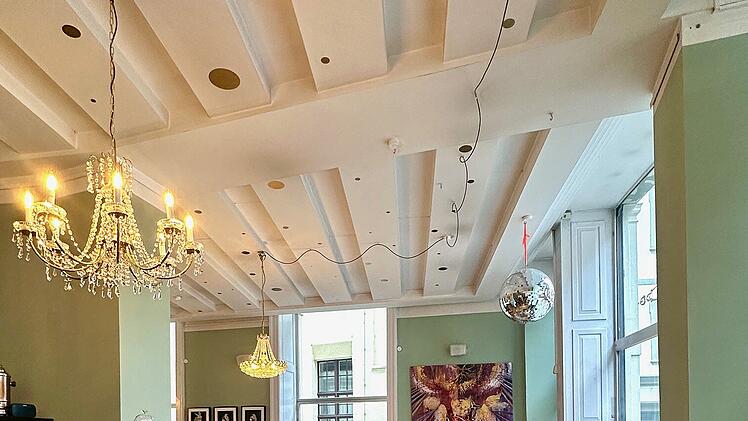 Ein stilvolles Caf&eacute; mit h&ouml;lzernem Boden, gr&uuml;nen W&auml;nden, eleganten Kronleuchtern und gem&uuml;tlichen Sitzgelegenheiten. Ein Kunstwerk und eine Diskokugel schm&uuml;cken den Raum.