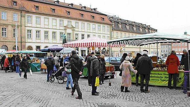 Auf dem Bild ist ein belebter Markt auf einem gepflasterten Platz zu sehen, mit mehreren St&auml;nden unter gestreiften Markisen. Menschen schlendern und betrachten die Produkte.