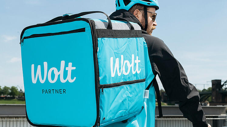 Eine Person tr&auml;gt eine gro&szlig;e, blaue Wolt-Liefertasche auf dem R&uuml;cken. Sie f&auml;hrt Fahrrad und tr&auml;gt einen blauen Helm.