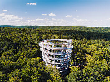 Ein spiralf&ouml;rmiger Turm ragt aus einem dichten, gr&uuml;nen Wald. Der Himmel ist klar und blau. Besucher spazieren auf dem Turm, der Teil des Baumwipfelpfads Steigerwald ist.