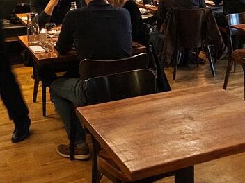 Ein Restaurant mit Holzst&uuml;hlen und -tischen. Menschen sitzen und unterhalten sich, ein Kellner bewegt sich schnell. Szenario erinnert an Zivilcourage bei einem medizinischen Notfall.