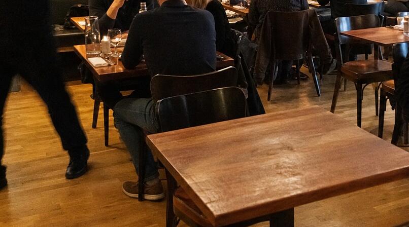 Ein Restaurant mit Holzst&uuml;hlen und -tischen. Menschen sitzen und unterhalten sich, ein Kellner bewegt sich schnell. Szenario erinnert an Zivilcourage bei einem medizinischen Notfall.
