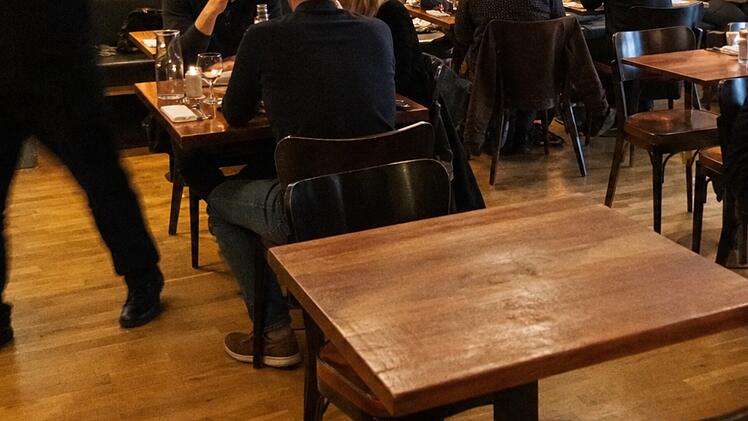 Ein Restaurant mit Holzst&uuml;hlen und -tischen. Menschen sitzen und unterhalten sich, ein Kellner bewegt sich schnell. Szenario erinnert an Zivilcourage bei einem medizinischen Notfall.