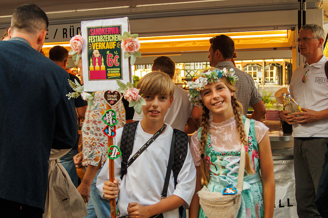 Zwei Kinder in Trachten halten ein Schild f&uuml;r den Festabzeichenverkauf auf der Bamberger Sandkerwa 2026. Menschen im Hintergrund und Getr&auml;nkeverkauf sichtbar.