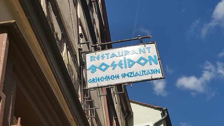 Ein Schild mit der Aufschrift "Restaurant Poseidon - Griechische Spezialitäten" hängt an der Hauswand. Der Himmel ist blau mit einigen Wolken.